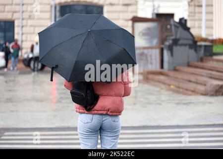 Pioggia in città, donna snella con ombrello che indossa jeans e giacca in piedi sulla strada а su sfondo crosswalk. Tempo piovoso in autunno Foto Stock