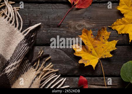 la plaid marrone di lana in una gabbia giace su un vecchio pavimento di legno come sfondo, uno sfondo di legno e su di esso una coperta e foglie autunnali, umore autunnale Foto Stock