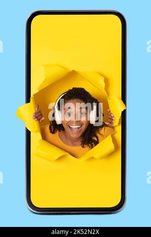 African Lady in smartphone indossando cuffie guardando attraverso il foro di carta Foto Stock