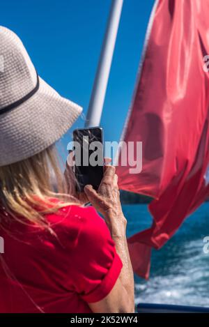 Donna bionda con berretto bianco e t-shirt rossa che scatta una foto con lo smartphone su una barca verso la bandiera svizzera. Foto Stock