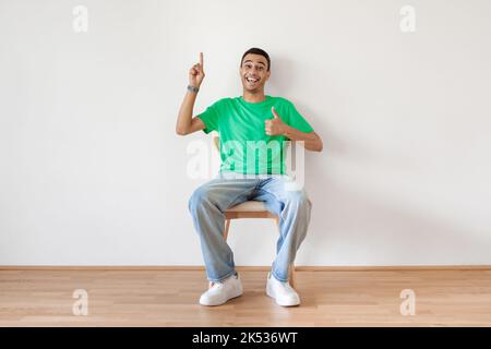Un uomo arabo allegro che gestiva i pollici verso l'alto e punta il dito verso l'alto, seduto sulla sedia sopra la parete bianca e sorridente alla macchina fotografica Foto Stock