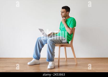 Uomo arabo travagliato seduto sulla sedia con il laptop, pensando alla soluzione al problema contro il muro bianco, copia spazio Foto Stock