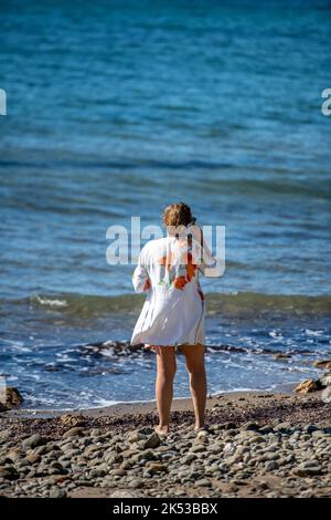 donna anziana che cammina lungo la spiaggia di sabbia utilizzando smartphone per fare una chiamata durante le vacanze sull'isola greca di zante o zante. Foto Stock
