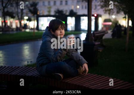 Un adolescente solitario con emozioni premurose e una tazza di carta in mano sulla strada in una serata autunno scuro sullo sfondo delle luci della città. Foto Stock