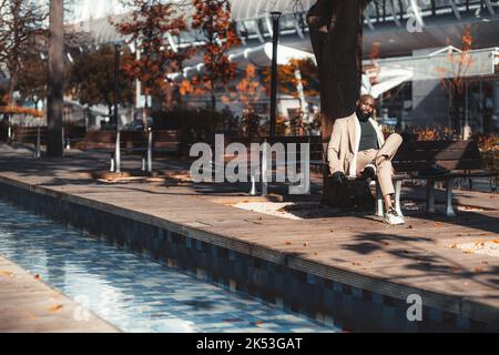 Un elegante imprenditore afro-americano calvo con una bella barba nera e un costume beige personalizzato è seduto su una panca di legno in un parco pubblico Foto Stock