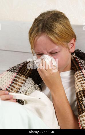 La bambina malata si avvolse in una coperta, guarda il termometro, soffia il naso in un tovagliolo. Telaio verticale. Il concetto di salute e medicina. Foto Stock