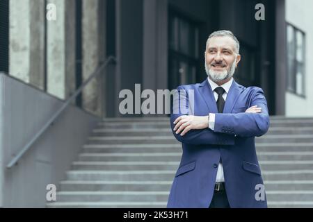 Ritratto dell'agente immobiliare senior bello uomo. Si trova accanto a un vestito da lavoro vicino a un edificio moderno, guarda di lato, sorride Foto Stock