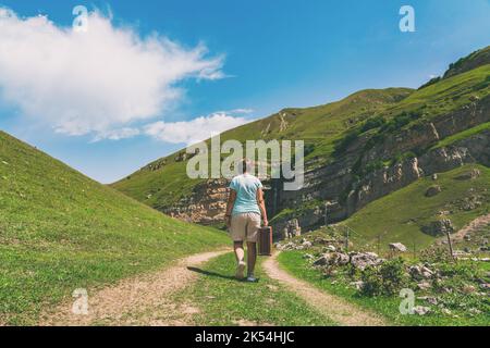 Una ragazza con una valigia cammina lungo una strada sterrata in montagna. Interessante viaggio della donna solitaria Foto Stock