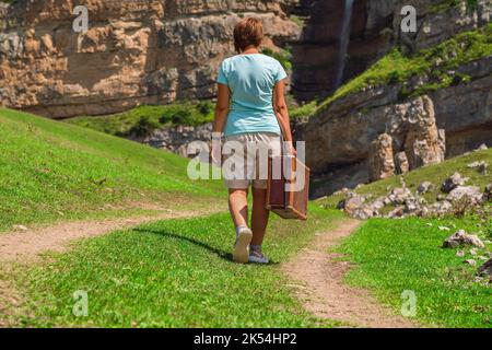 Una ragazza con una valigia cammina lungo una strada sterrata in montagna. Interessante viaggio della donna solitaria Foto Stock