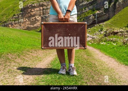 Una ragazza con una valigia cammina lungo una strada sterrata in montagna. Interessante viaggio della donna solitaria Foto Stock