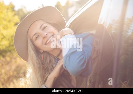 Sognando donna con gioia lasciare la città che si sta attaccando testa fuori dal finestrino dell'auto aperto. Giovane donna bionda in cappello con umore felice andare a viaggio in auto. In viaggio Foto Stock