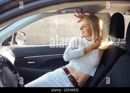 Donna attraente con gioia lasciare la città seduta in spazioso salone di auto nero. Giovane donna bionda con umore felice andare a viaggio in auto in giornata di sole. In viaggio Foto Stock