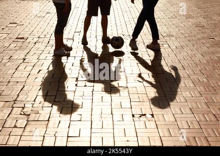 Divertimento tra amici. Immagine ritagliata delle gambe che calcia una palla con ombre sulla strada asfaltata. Foto Stock