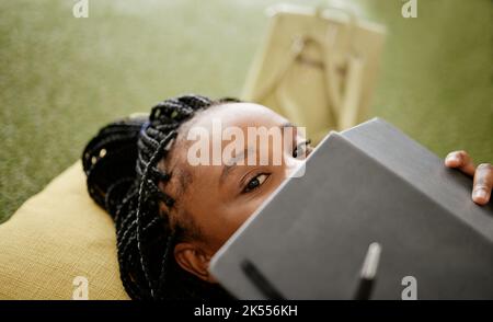 Donna nera e studente che si nasconde faccia con notebook su casual studio lounge borsa di fagioli. Timido studente universitario africano sullo studio pausa di riposo Foto Stock