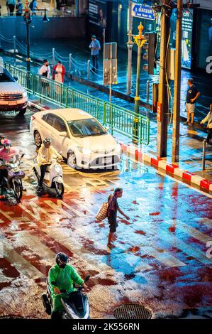 Un uomo tailandese a piedi nudi attraversa la zebra attraversando la famosa e trafficata Asoke montri Rd Incrocio stradale a Bangkok, Thailandia in una serata piovosa bagnata. Foto Stock