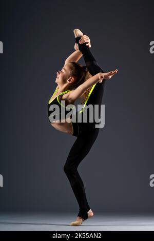 Giovane ginnastica. Una ragazza adolescente in una tuta è impegnata in ginnastica, facendo esercizi di stretching su uno sfondo grigio. Foto di alta qualità Foto Stock
