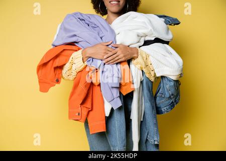 vista ritagliata di felice donna afroamericana che tiene un mucchio di vestiti colorati sul giallo, immagine di scorta Foto Stock