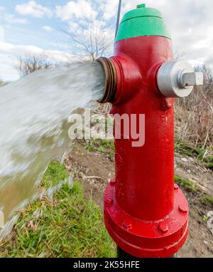 Acqua che fluisce da un idrante rosso fuoco aperto. Vista laterale primo piano. La parte superiore è verde. L'idrante viene bagnato dallo spray. Foto Stock