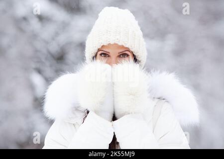 Primo piano ritratto di donna attraente in bianco cappello lavorato a maglia, e pellicce mitens su neve freddo inverno sfondo natura. Occhi marroni belli, modello di copertura Foto Stock