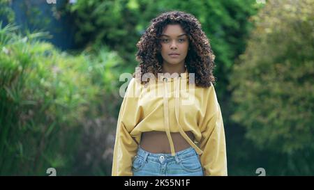 Una giovane ragazza nera ispanica latina 20s in piedi all'aperto guardando la macchina fotografica. Donna millenaria afroamericana con capelli ricci che indossa blusa gialla an Foto Stock