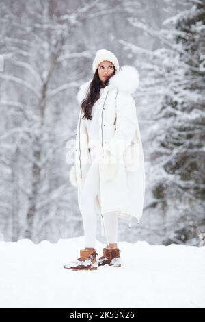 Ritratto esterno a tutto corpo di una giovane bella donna alla moda che indossa un elegante piumino bianco invernale, cappello a maglia, guanti, leggings. Posa del modello Foto Stock