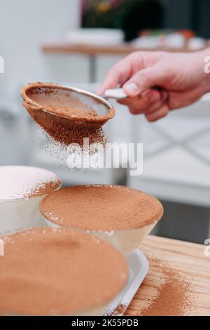 Il processo di preparazione del dessert tiramisù. Il piatto viene cosparso di cacao attraverso un setaccio. Primo piano, messa a fuoco selettiva. Foto Stock