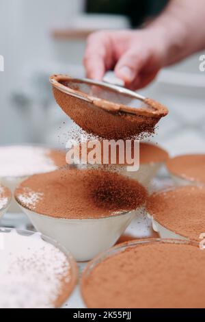 Il processo di preparazione del dessert tiramisù. Il piatto viene cosparso di cacao attraverso un setaccio. Primo piano, messa a fuoco selettiva. Foto Stock