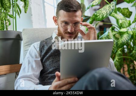 Giovane uomo orientale indiano rilassato e tranquillo seduto sul pullman nel moderno soggiorno con tablet computer controllare i social media, navigare in Internet Foto Stock