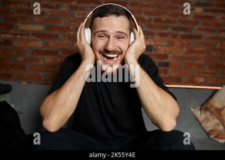 Bell'uomo bearded in cuffie che ascoltava la musica a casa. Musica. Ritratto a metà lunghezza di giovane ragazzo sorridente in abiti casual stile gioventù seduto Foto Stock