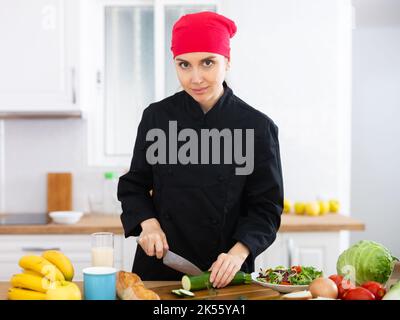 Sorridente cuoco femminile in uniforme nera che prepara insalata di verdure in cucina privata Foto Stock