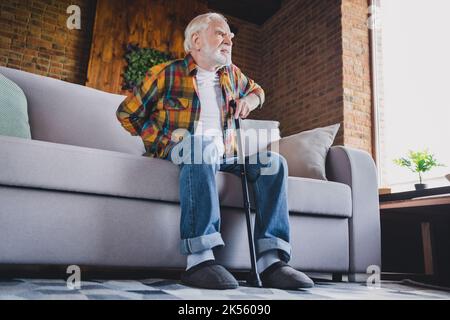 Foto di un uomo stanco infelice pensionato indossare camicia a scacchi cercando in piedi sul divano tenendo in mano canne da passeggio casa camera interna Foto Stock