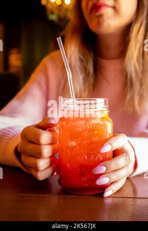 Le mani della giovane donna caucasica tengono il vetro con manico di limonata di fragola rossa con ghiaccio e paglia bevente sul tavolo di legno. Mani femminili con mani rosa Foto Stock