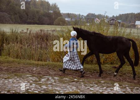 Una giovane donna vestita come una donna contadina europea del 17th ° secolo conduce un cavallo attraverso un campo in una serata d'autunno Foto Stock