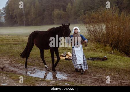 Una giovane donna vestita come una donna contadina europea del 17th ° secolo conduce un cavallo attraverso un campo in una serata d'autunno Foto Stock