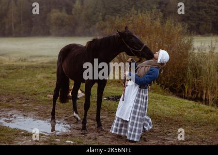 Una giovane donna vestita come una donna contadina europea del 17th ° secolo conduce un cavallo attraverso un campo in una serata d'autunno Foto Stock