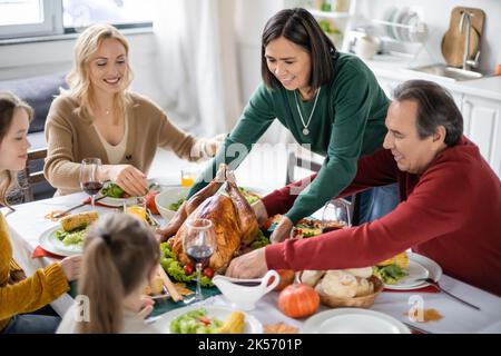 Donna multiculturale mettendo tacchino sul tavolo vicino a cena di ringraziamento e famiglia a casa, immagine stock Foto Stock