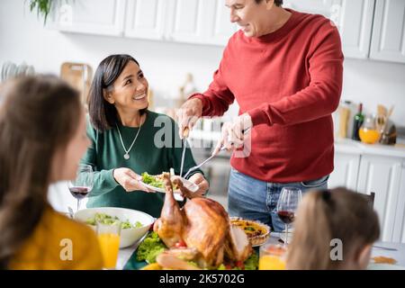 L'uomo anziano che mette il tacchino sulla piastra vicino alla famiglia interracial che celebra il ringraziamento nel paese, immagine di riserva Foto Stock