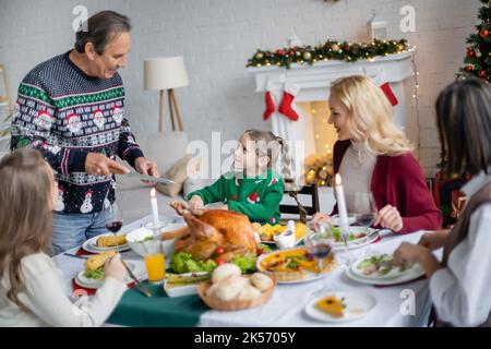 ragazza sorridente guardando nonno con coltello mentre avendo cena di natale con la famiglia interracial, immagine di scorta Foto Stock