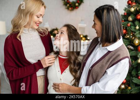 ragazza gioiosa guardando madre sorridente vicino multirazziale e decorato albero di natale, immagine stock Foto Stock