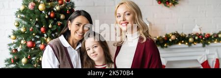 ragazza felice con madre e multirazziale che guarda la macchina fotografica in soggiorno con decorazione di natale, banner, immagine stock Foto Stock