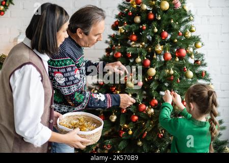 donna multirazziale che tiene il cestino di vimini con i baubles vicino al marito e alla nipote che decorano l'albero di natale, immagine di riserva Foto Stock