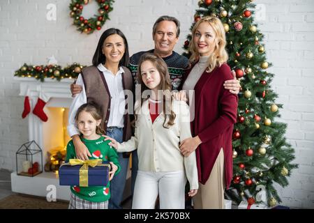 sorelle felici con nonni interracial e madre che guardano la macchina fotografica in soggiorno con la decorazione di natale, immagine di riserva Foto Stock