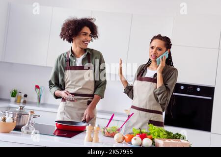 Foto di arrabbiato moglie marito sposo vestito grembiule cucina pranzo parlando moderno gadget in casa camera Foto Stock