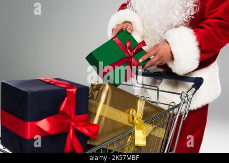 Vista ritagliata di babbo natale in costume mettere scatola regalo nel carrello isolato su grigio, immagine stock Foto Stock