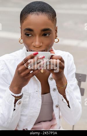 Ritratto di elegante donna afroamericana in giacca bianca bere caffè su strada urbana, immagine stock Foto Stock