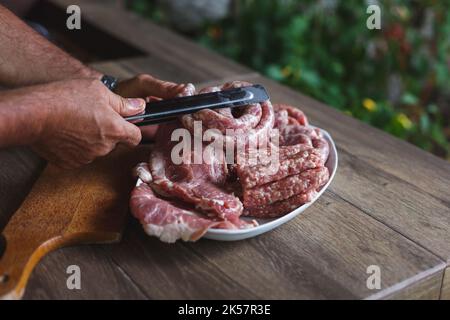 Chef maschile con pinze che tiene in mano salsicce appena preparate, pronte per il barbecue. Messa a fuoco selettiva, spazio di copia Foto Stock