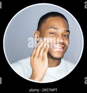 Cura della pelle dell'uomo. Prodotto cosmetico. Cura del corpo. Ritratto di un ragazzo africano soddisfatto che applica crema viso su pelle scura sorridendo isolato sul dorso grigio Foto Stock