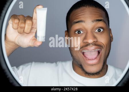 Cura della pelle maschile. Pubblicità di prodotti cosmetici. Idratante per la pelle. Sorpreso uomo africano che mostra bianco mockup viso crema tubo isolato su grigio Foto Stock