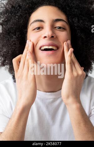 Cura del viso dell'uomo. Trattamento della pelle. Trattamento dell'acne. Felice soddisfatto bel ragazzo con lunghi capelli ricci marrone in t-shirt bianca applicando crema godendo touc Foto Stock