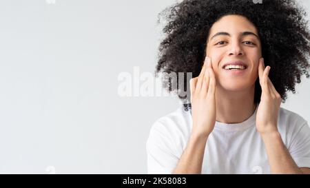 Cura della pelle dell'uomo. Cosmetologia facciale. Felice soddisfatto bel ragazzo con lunghi capelli castani ricci applicando crema godendo toccare morbido viso sorridente isolato Foto Stock
